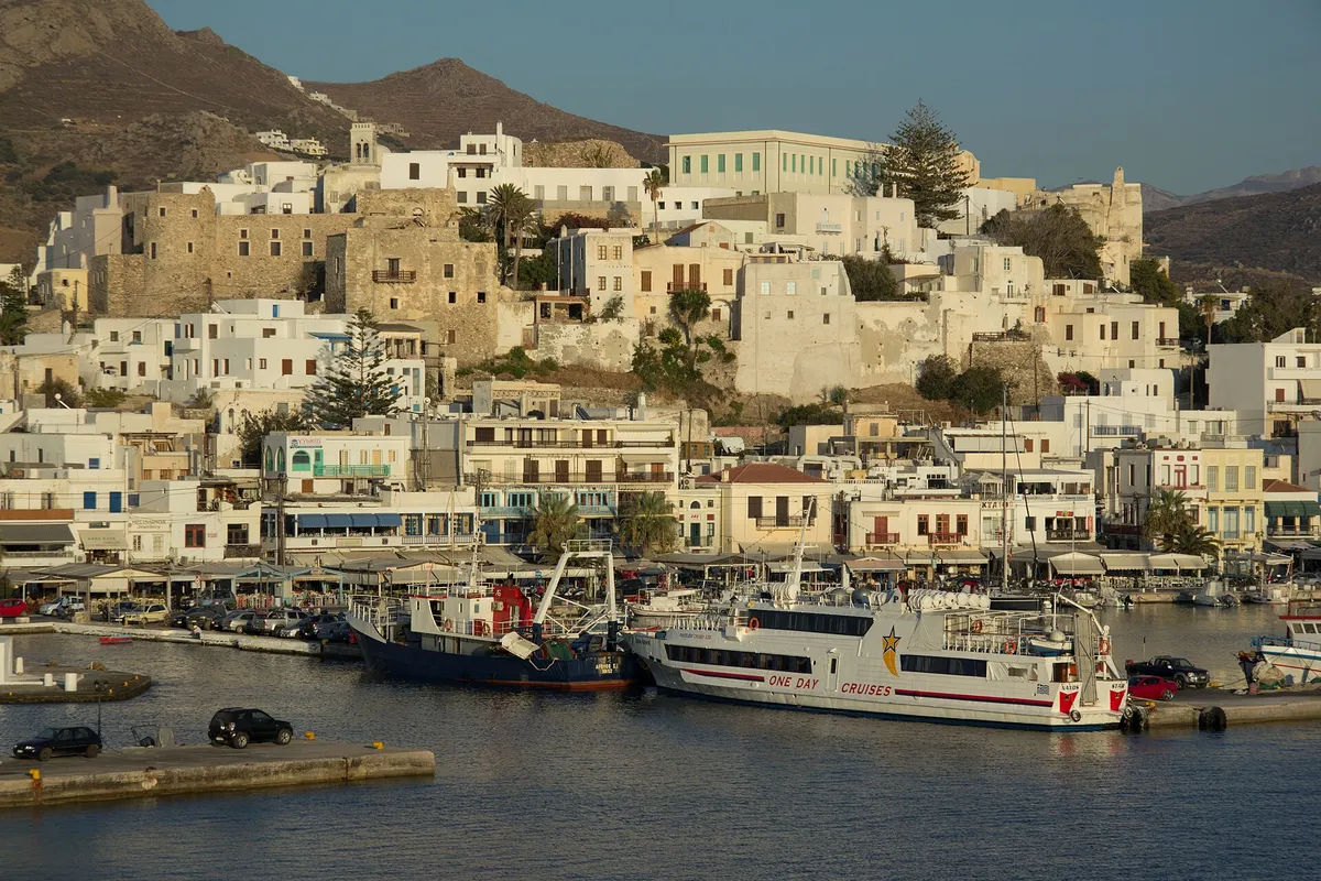 Naxos town waterfront at golden hour with the Kastro hill and a day-cruise boat moored in the port