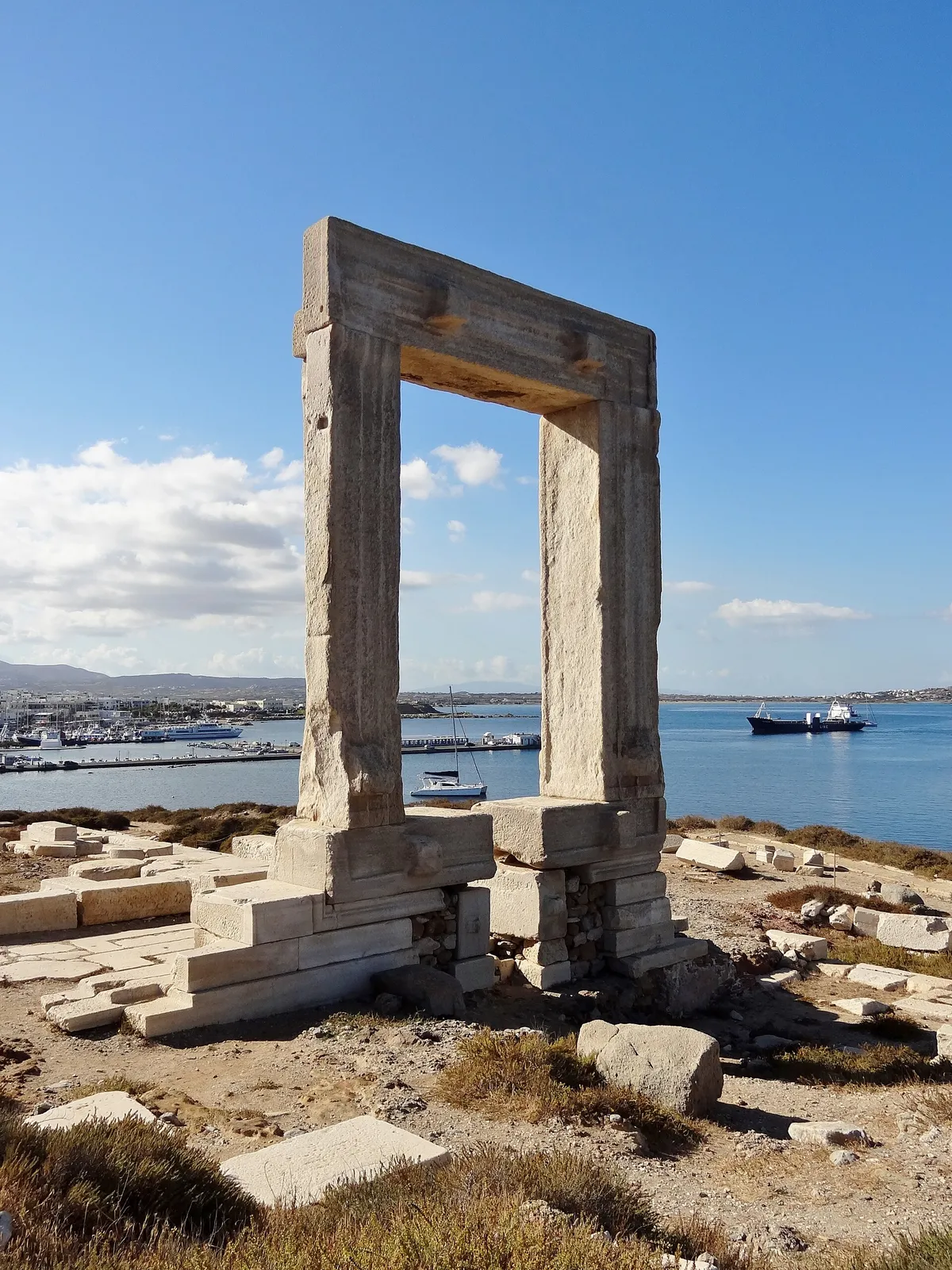 The Portara marble gate of Naxos with the harbour and boats behind under a blue sky