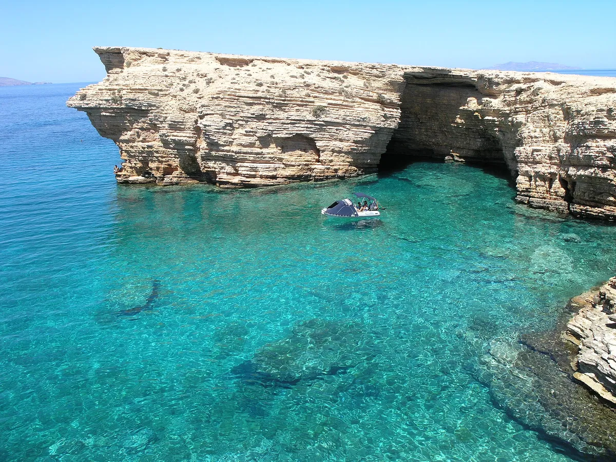 Small boat at anchor beneath a golden sea arch and cliffs in turquoise Koufonisia water near Naxos, Greece